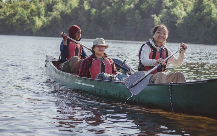 three people wearing life jackets paddle a canoe toward the camera. The person in the front of a canoe smiles at the camera. 